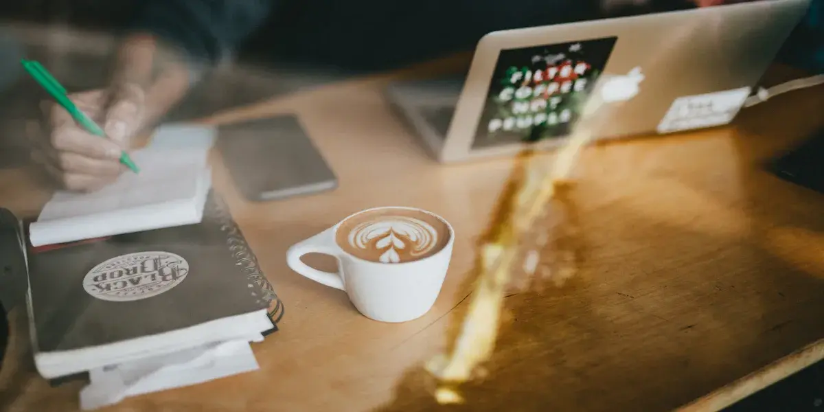 Mano escribiendo en una libreta junto a una taza de café con leche